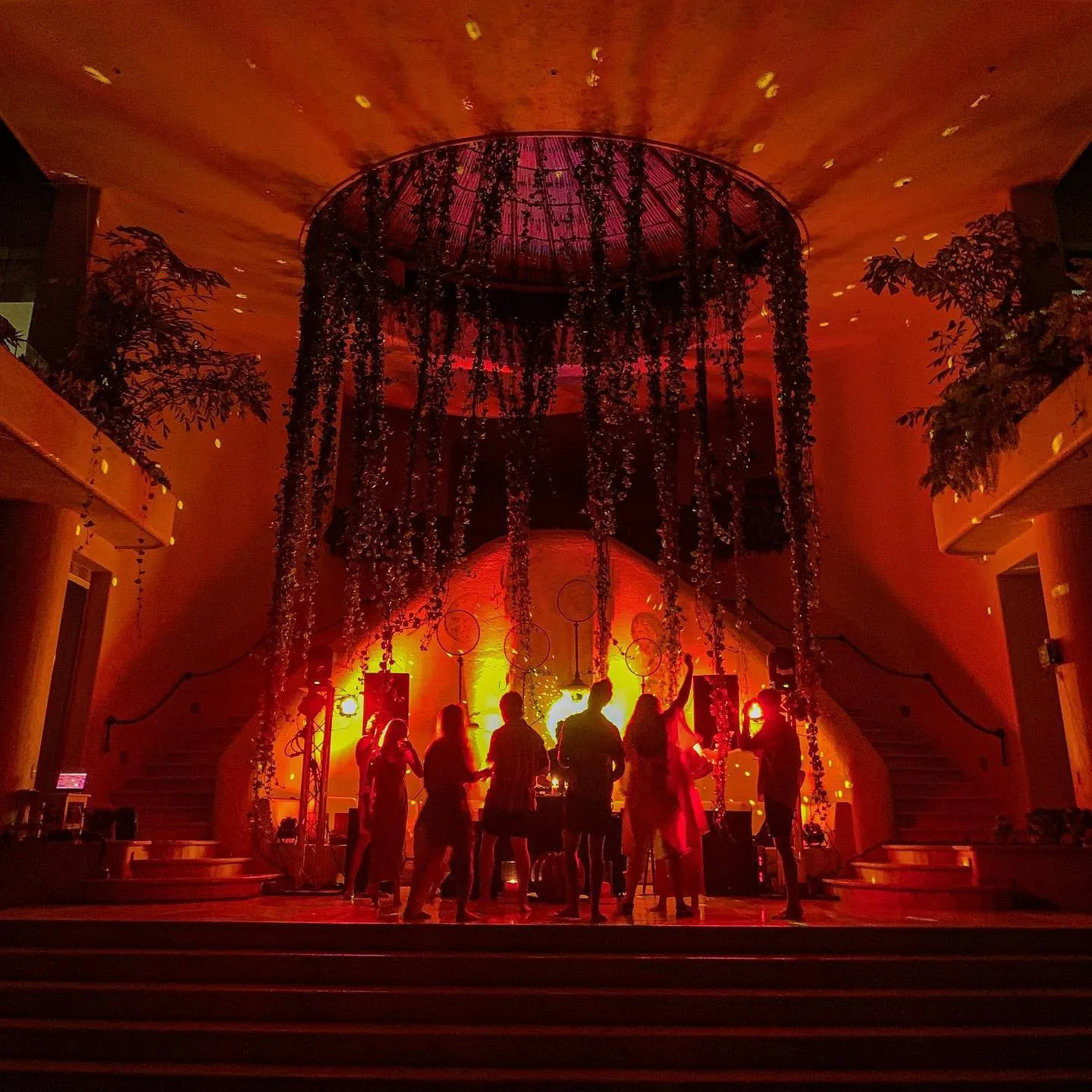 A courtyard lit red at night, with hanging floral installation and uplit trees